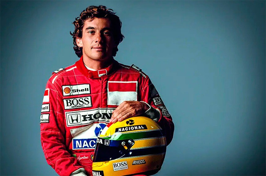 Ayrton Senna de macacão vermelho, um capacete de corrida amarelo em seu colo e sorrindo para a câmera