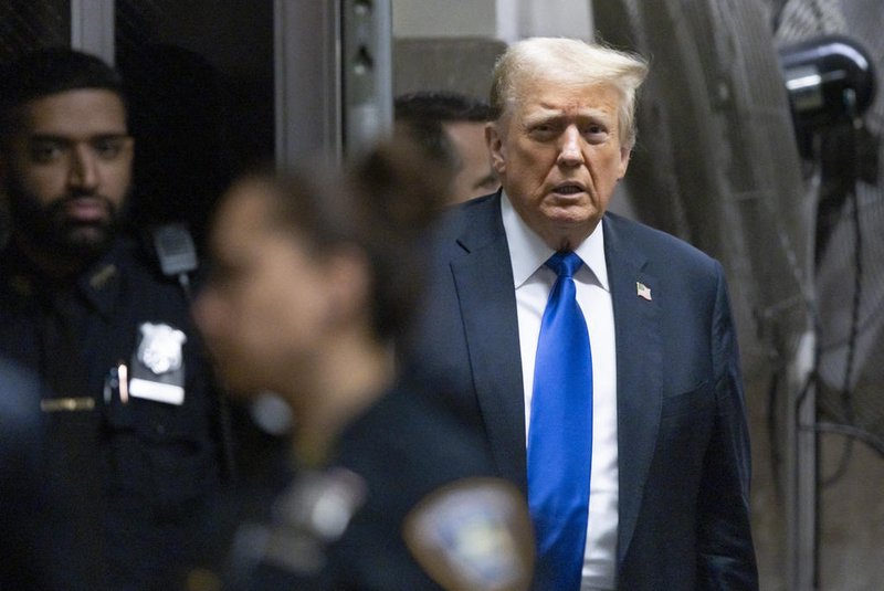 Donald Trump de terno preto, gravata azul e blusa branca, caminhando na rua